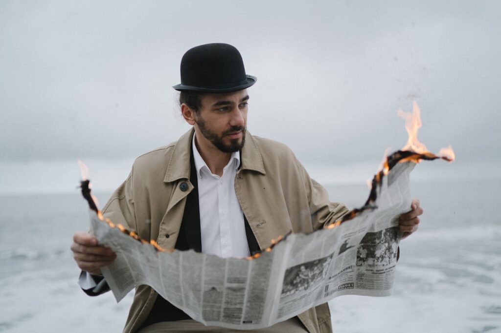 A man in a bowler hat and coat reads a burning newspaper by the ocean on a cloudy day.